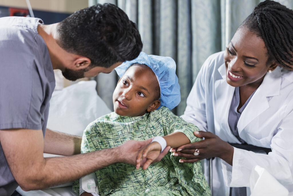 En 6-årig afrikansk-amerikansk pige, der sidder på en hospitalsseng med en læge og sygeplejerske, der undersøger hende.  Hun har en hospitalskittel og kirurgisk hætte på. En sygeplejerske i kittel, en ung mellemøstlig mand i 20’erne, står på den ene side af hende, og en kvindelig læge, en moden afrikansk-amerikansk kvinde i 40’erne, står på den anden side.