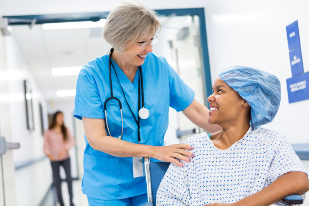 Ældre voksen hvid kvindelig sygeplejerske med gråt hår skubber patient i kørestol. Hun tager patienten med til opvågning efter ambulant kirurgisk procedure. Patienten er en ung voksen afrikansk-amerikansk kvinde, som har en hospitalskittel og kirurgisk hætte på. Kvinderne smiler, mens de taler med hinanden.